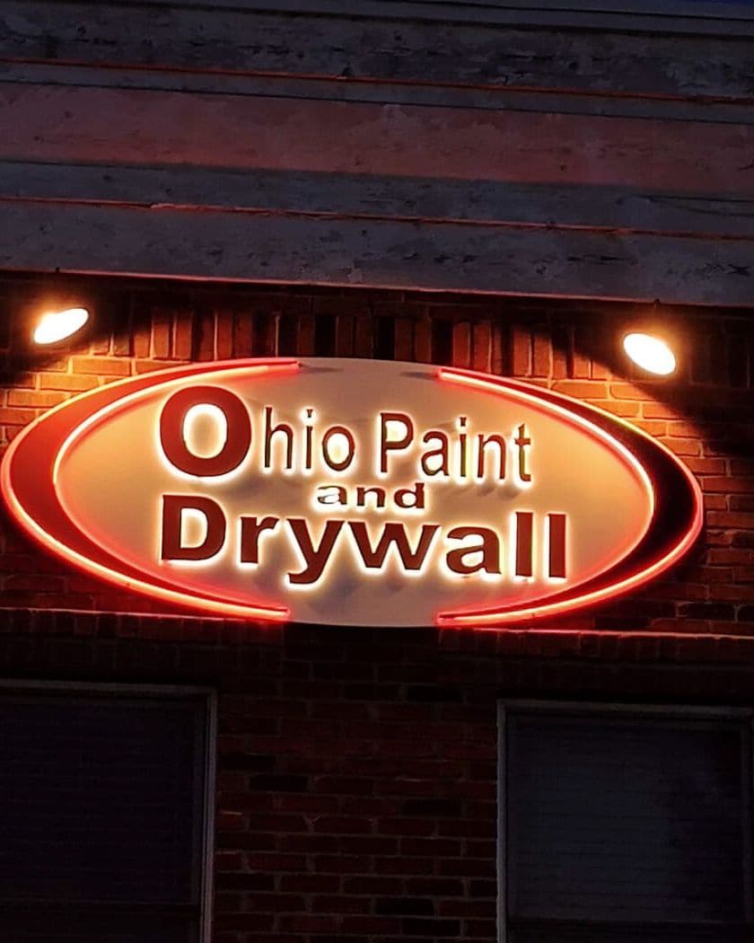 Lit oval Ohio Paint and Drywall sign glowing red on the brick front of the Waterville office at night